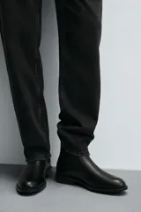 Bota estilo Chelsea de cuero negro con diseño liso, paneles elásticos laterales y tirador trasero. Presenta horma redonda y suela con tacón bajo.