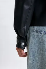 Sobrecamisa de cuero sintético en color negro, con cuello camisero, cierre frontal con botones metálicos a presión y mangas largas con puños abotonados.