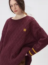 Sweater de punto grueso color bordó con diseño de rombos en relieve. Presenta cuello redondo, corte holgado, aberturas laterales y puños con rayas amarillas en contraste. Incluye logo bordado en el pecho.