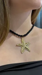 Gargantilla tipo choker con cordón trenzado negro y dije grande de estrella de mar en tono dorado con textura granulada.