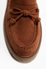 Mocasines de gamuza color marrón con detalle de tiras anudadas en el empeine, costuras decorativas y suela track gruesa de goma.