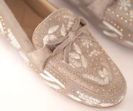 Mocasín de gamuza color avena con bordados florales en hilo blanco y pequeños apliques metálicos dorados. Presenta un lazo decorativo en la capellada y suela de goma flexible.