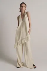 Top halter color crudo, de escote en V y diseño confeccionado en hilatura con mezcla de viscosa. Presenta bajo asimétrico con flecos al tono y cierre en la espalda.