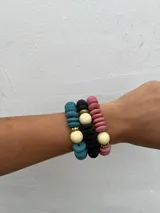 Set de tres pulseras elásticas con cuentas de madera en colores celeste, negro y rosa, con detalles de cuentas doradas y cuentas color crema.