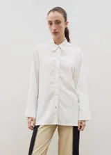 Camisa de corte holgado y diseño básico, confeccionada en color blanco con costuras visibles en contraste. Presenta cuello clásico, cierre frontal con botones y puños anchos.