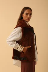 Chaleco de piel sintética en color chocolate, con cierre frontal y detalles de cuero en los bordes. Presenta un diseño de cuello solapa y ajuste lateral con hebillas.