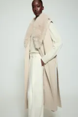 Chaleco largo de mezcla de lana color beige, de corte recto con manga sisa. Cuenta con cuello solapa con aplicación de pelo sintético extraíble, bolsillos laterales ocultos y un cinturón ajustable del mismo tejido para anudar en la cintura.