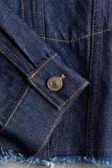 Campera cropped de denim de algodón rígido, color azul, con cuello, cierre frontal de botones y hombros caídos. Cuenta con bolsillos superiores con solapa y botón, puños abotonados y dobladillo deshilachado.