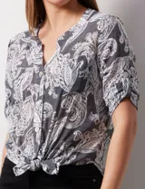 Blusa celeste con estampado blanco, de gasa, con cuello a la base y escote en V. Tiene abertura con botones, bolsillos delanteros y mangas 3/4 regulables con presilla y botón. El ruedo termina con un nudo.