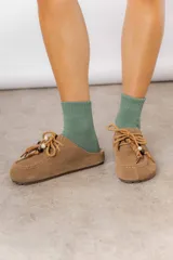 Zueco de cuero gamuzado color tostado, con cordones decorativos adornados con cuentas de madera y piezas envueltas en cuero. Diseño tipo mocasín con costuras visibles en la parte superior y abierto en la parte trasera.