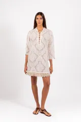 Vestido camisero corto de encaje blanco con diseño ornamental, escote en V con cordón ajustable y ojales metálicos, mangas 3/4 acampanadas y ruedo con detalle de flecos de caracoles tejidos a mano.