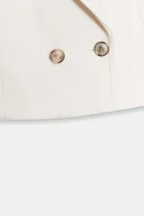 Blazer corto blanco de cuello solapa y manga larga con hombreras. Presenta un cierre frontal cruzado con dos botones dorados.