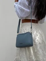 Bandolera pequeña de diseño rectangular en color beige, confeccionada en cuero sintético. Presenta una correa larga ajustable del mismo material y cierre superior con cremallera. Incluye el logo de la marca grabado en la parte frontal.