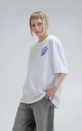 Remera blanca de corte holgado con estampa central en color azul que combina motivos geométricos tipo chakana con figuras de aves dispuestas en círculo.