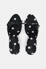 Sandalia tipo mule negra con estampado de lunares blancos, efecto satinado, punta fina, tacón fino y tira delantera con lazo decorativo.