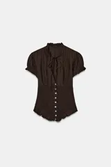 Top de color marrón con textura acanalada, cuello redondo con lazada y escote en pico. Presenta mangas cortas abullonadas con terminación elástica, cuerpo con tejido tipo nido de abeja y cierre frontal con botones.