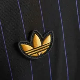 Camiseta de fútbol Adidas del Manchester United, modelo Terrace Icons. Diseño negro con finas rayas verticales en violeta, detalles dorados en las tres tiras de los hombros, logo de Adidas y escudo del club. Cuello redondo con ribete violeta y amarillo.