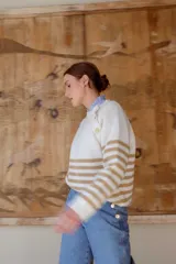 Sweater de punto en color crema con rayas horizontales en tono dorado. Presenta un diseño de cuello redondo con una solapa lateral decorada con botones metálicos dorados y corte holgado.