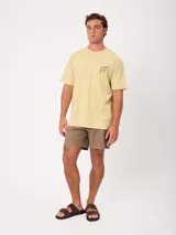 Remera de manga corta color amarillo, corte holgado, con gran estampado en la espalda de un perro en bicicleta con una tabla de surf y un loro, acompañado del texto "Wild by Nature Rusty Worldwide".