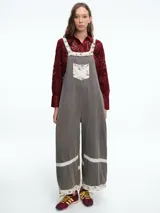 Mono de corte holgado en color gris, con tirantes ajustables y bolsillo frontal tipo parche. Presenta detalles decorativos de encaje y tela estampada en los bordes de los tirantes, el bolsillo, y el ruedo de las piernas.