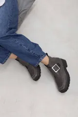 Botines de cuero vacuno color suela con textura envejecida, suela track negra con pespunte en contraste, solapa cruzada con gran hebilla metálica plateada y cierre lateral interior.