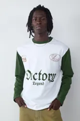 Remera de manga larga con diseño estilo camiseta de fútbol, cuerpo blanco y mangas verdes. Presenta estampados gráficos en el frente con la inscripción 'Victory Legend', el número '39' y un emblema ovalado.