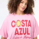 Camiseta oversize de manga corta color rosado, con cuello redondo y estampa frontal con la leyenda "Costa Azul" en rojo y amarillo.