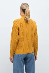 Polera de tejido de punto color ocre con cuello alto, diseño de rombos en relieve en el frente y mangas con detalle de trenzas laterales.