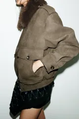 Cazadora color verde militar con efecto ante, cuello solapa con aplicación de pelo sintético desmontable, manga larga con puño, trabillas en los hombros y bolsillos frontales con solapa y botón. Cierre frontal con cremallera y botones ocultos.