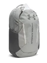 Mochila Under Armour modelo Hustle 6.0, color gris, con tecnología UA Storm resistente al agua. Cuenta con compartimento acolchado para portátil, bolsillo para calzado o ropa sucia, bolsillos laterales de malla y sistema Molle para sujeción.