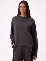 Sweater de corte oversize y estilo cropped, color gris oscuro, con cuello redondo y detalle de costura en forma de X en el pecho. Presenta un pequeño logo bordado en el ruedo inferior.