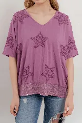 Blusa lila de algodón con escote en V, mangas cortas y holgadas. Presenta estrellas bordadas en relieve en la parte superior y un borde de lentejuelas en el dobladillo inferior.
