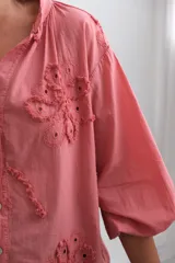 Blusa color coral de corte holgado, con cuello mao y cierre frontal de botones. Presenta mangas largas abullonadas y detalles de flores bordadas con técnica de calado en el frente.