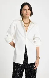 Camisa blanca de corte recto con cuello clásico estructurado, mangas largas con puños ajustados y cierre frontal de botones ocultos. Presenta un panel trasero con frunces y detalle elástico, con un bajo ligeramente redondeado.