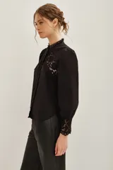 Camisa negra de algodón con detalles de encaje calado en el pecho, hombros y puños. Presenta cuello alto con terminación de encaje, cierre frontal con botones y mangas largas abullonadas.