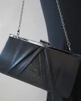 Clutch de mano color negro con diseño estructurado, detalles de pliegues y panel central con incrustaciones de cristales brillantes. Incluye marco metálico plateado y cadena desmontable para usar al hombro.