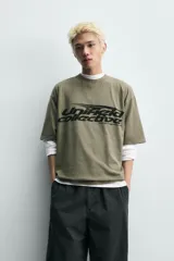 Camiseta de algodón con corte relajado, cuello redondo y manga corta. Presenta un estampado gráfico frontal en color negro con tipografía estilo deportivo y efecto desgastado.