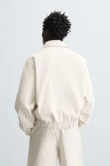 Cazadora de hombre color beige claro, de corte relajado, con cuello solapa, cierre frontal de cremallera y puños y bajo elásticos. Presenta una textura de rayas sutiles y efecto lavado.