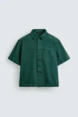 Camisa de denim de algodón color verde oscuro, de corte relajado (relaxed fit) y manga corta. Posee cuello solapa, bolsillo plastrón en el pecho con bordado a tono y cierre frontal de botones.