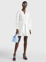 Vestido camisero corto color blanco, confeccionado en algodón orgánico. Presenta cuello en V, cierre frontal con botones, mangas largas abullonadas, cinturón de lazo ajustable en la cintura, bolsillos laterales y detalle de encaje calado en forma de escalera.