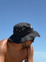 Gorro tipo bucket negro con cordón regulable para ajuste. Presenta un pequeño logo blanco en el frente.