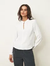 Blusa de manga larga color blanco, con cuello mao y abertura frontal tipo gota con cierre de botón. Presenta un diseño fluido con frunces en el escote y puños abotonados.