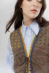 Chaleco de algodón color marrón con bordados decorativos en tonos azul, amarillo y fucsia. Presenta un diseño corto con cierre frontal mediante tiras para atar y detalles de borlas.