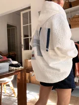 Campera tipo hoodie oversize de textura tipo corderito en color blanco, con diseño de parches en la espalda que combinan tela de jean a rayas y encaje blanco. Cuenta con capucha y un pequeño parche de cuero sintético en la parte inferior trasera.