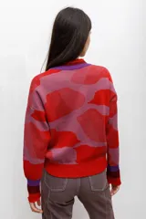 Buzo tejido en técnica jacquard con diseño de formas abstractas en tonos rojos, rosados y violetas. Presenta cuello redondo, mangas largas y puños y ruedo acanalados.