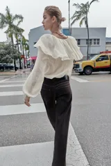 Blusa de color beige con escote en pico y lazo frontal decorado con abalorios. Presenta mangas largas con puños elásticos y un detalle de volantes amplios con terminación de puntilla a tono sobre los hombros y el pecho.