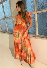 Vestido largo naranja con estampado geométrico y paisley, escote en V con bordado colorido, mangas cortas con volados y falda amplia en capas con cordón ajustable terminado en borlas turquesas.