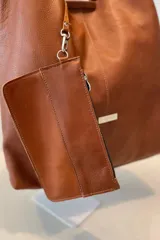 Bolso tipo tote de gran capacidad, confeccionado artesanalmente en cuero vacuno color marrón. Presenta un diseño minimalista con costura central vertical, fuelle lateral ajustable con cierre y placa metálica con logo en la parte inferior.