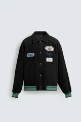 Cazadora varsity de pana de algodón ligeramente acolchada, con cuello solapa, cierre frontal con botones a presión y puños y bajo con acabado en rib a rayas. Presenta múltiples parches bordados decorativos en el frente y bolsillos laterales de vivo.
