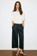 Pantalón de popelina de tiro alto y pernera ancha, de color negro. Presenta pinzas delanteras, bolsillos laterales y falsos bolsillos de vivo en la espalda. Incluye un cinturón combinado tipo pañuelo trenzado con tiras colgantes y cuentas de madera. Cierre frontal con cremallera, botón interior y gancho.